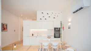 A modern kitchen with white cabinets, a dining area with a wooden table and chairs, and a small seating area with a blue chair and a patterned cushion. - Liiiving in Porto - Haven Pool Apartment (Porto)