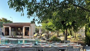 Un espace aménagé au bord de la piscine, avec des tables et des chaises en bois, un petit bâtiment doté d’un toit en tuiles, et un jardin où poussent différentes plantes et un arbre.