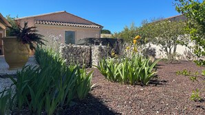 Une maison avec un toit en tuiles, un mur en pierre et un jardin agrémenté de plantes et d’un grand bac à fleurs.