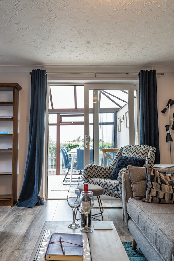 Ein Wohnzimmer mit einer Couch, einem Tisch mit Buch und Gläsern, einem Sessel und Blick durch eine Glastür in einen Außenbereich.