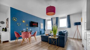 A modern living room with a blue accent wall, a flat-screen TV, a dining area with pink chairs, and a blue sofa. - The Post House Two by Fortuna Property Portfolio, The Post House Two Apartment (Gloucester District)
