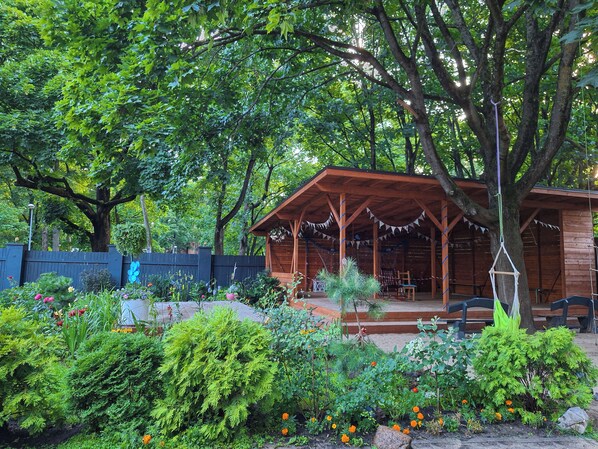 A wooden gazebo with a covered porch, surrounded by lush greenery and flowering plants.