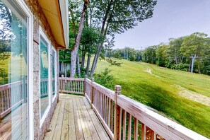 A wooden deck with glass railings overlooking a grassy area with trees and a distant view of a ski lift.