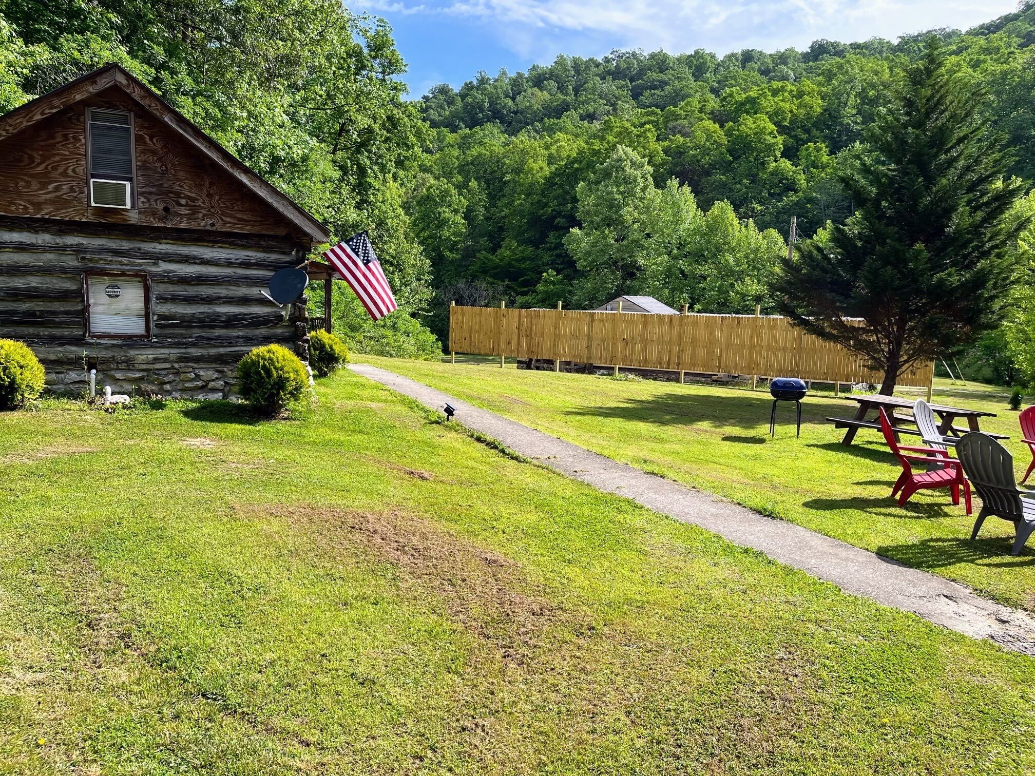 A rustic cabin with a stone foundation, an American flag, and a picnic area with chairs and a table.