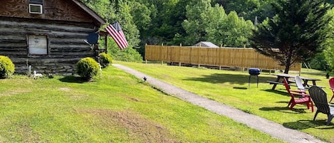 A rustic cabin with a stone foundation, an American flag, and a picnic area with chairs and a table.