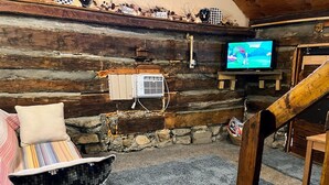 A cozy log cabin interior with a television, a stone wall, and a wooden staircase. - Quaint old fashioned cabin, Loft, Grill, Fire Pit!  (Stanton)