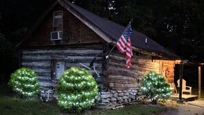 A rustic log cabin with an American flag, two illuminated trees, and a stone wall. - Quaint old fashioned cabin, Loft, Grill, Fire Pit!  (Stanton)