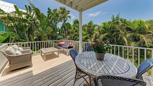 Une terrasse extérieure couverte, dotée d’une table et de chaises en bois, entourée d’une végétation tropicale luxuriante et d’une balustrade blanche.