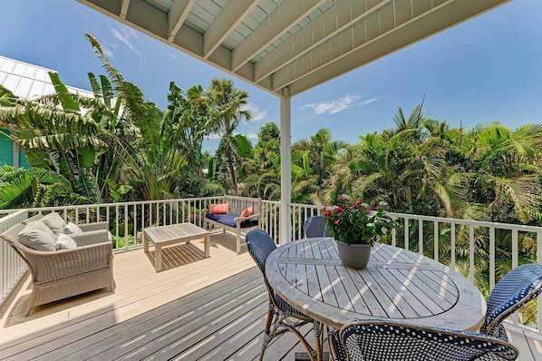 Una terraza cubierta al aire libre con una mesa y sillas de madera, rodeada de exuberante vegetación tropical y una barandilla blanca.