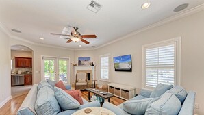Un vaste salon doté d’un ventilateur de plafond, d’une cheminée, d’un téléviseur et offrant une vue sur l’extérieur grâce à de grandes fenêtres.