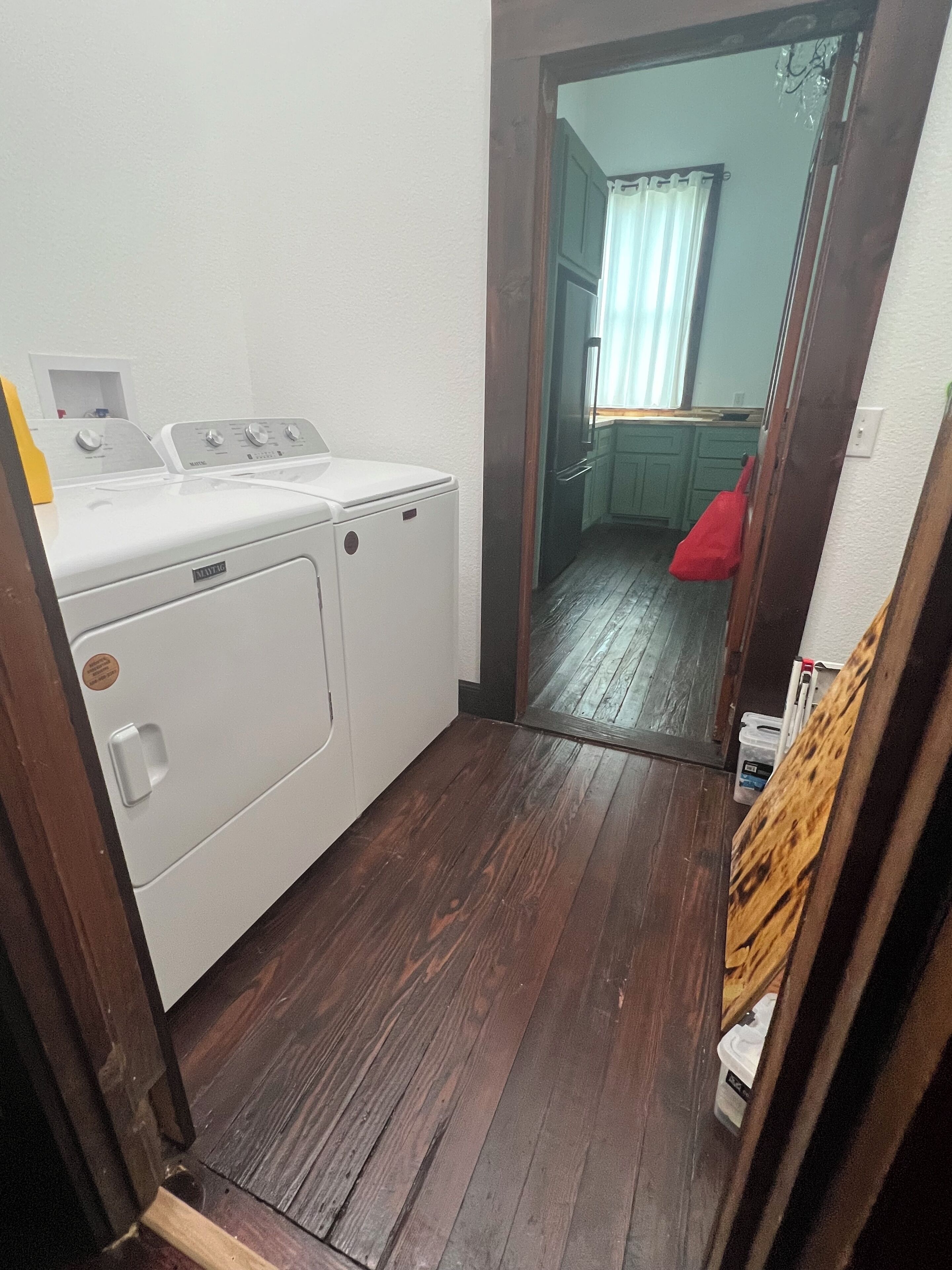 Un cuarto de lavandería con lavadora y secadora blancas, piso de madera y un acceso a un área de cocina.