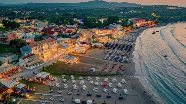 An aerial view of a coastal town with a beach lined with rows of sunbeds and boats, surrounded by buildings and a hilly landscape in the background.