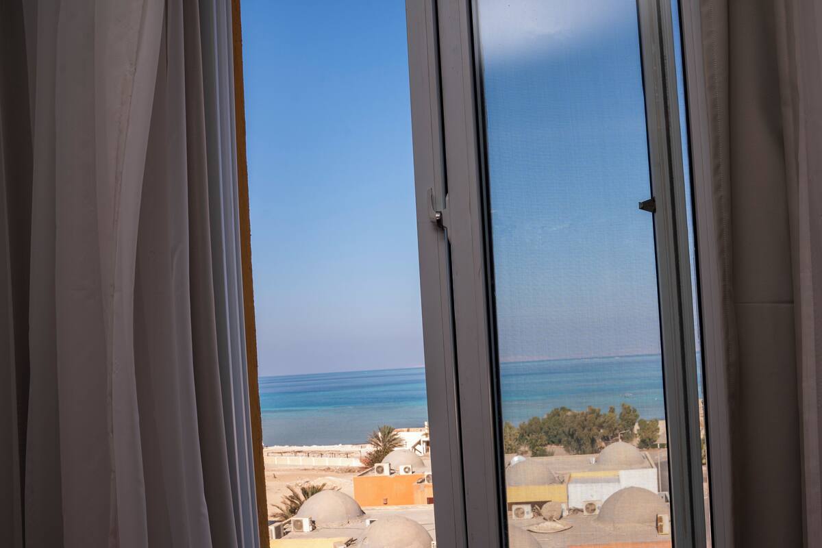 A view through a window with white curtains, revealing a coastal landscape with buildings and a clear sky.