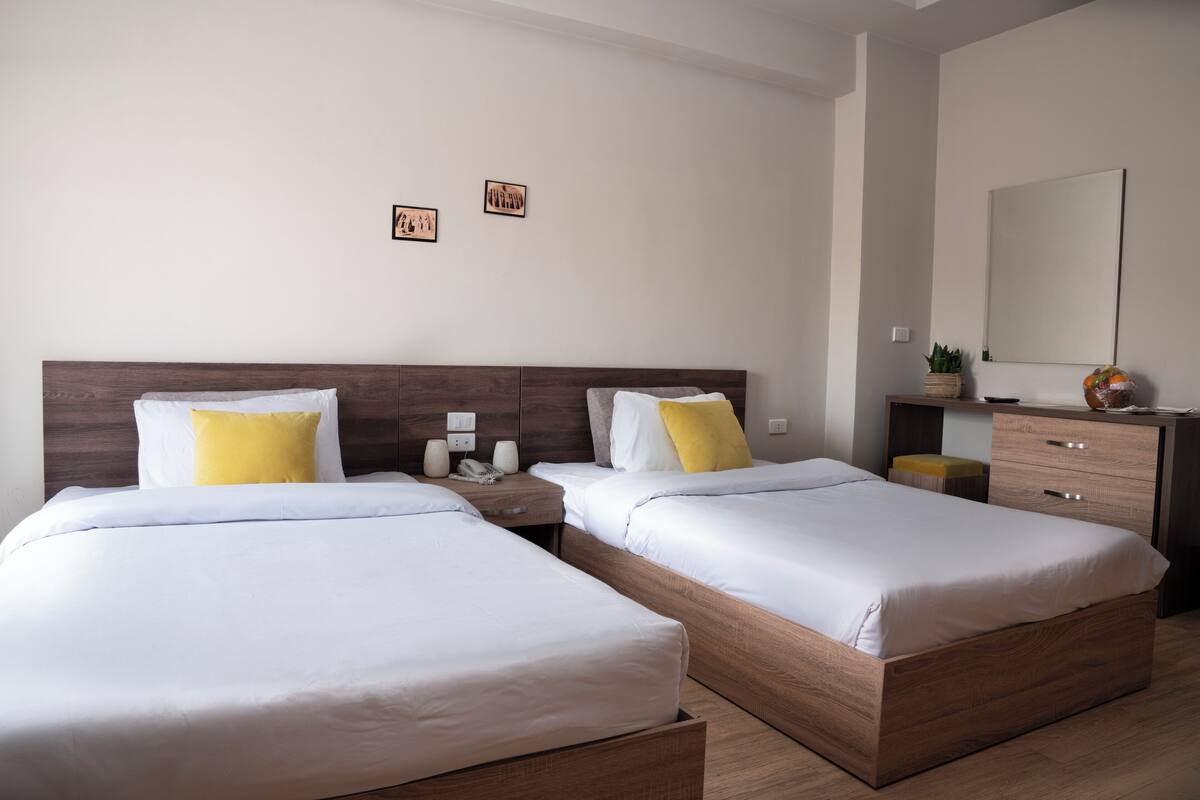 A hotel room with two beds, a wooden headboard, a nightstand, a mirror, and a small plant.