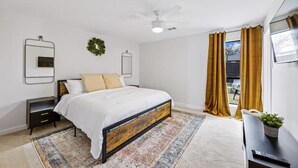 Un dormitorio con cama, mesitas de noche, ventilador de techo, ventana con cortinas y una pequeña mesa con una planta.