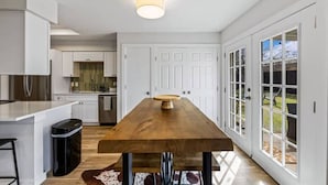 Cocina moderna con mesa de comedor de madera, muebles de color blanco y vistas al exterior a través de grandes ventanales.