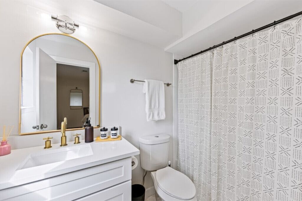 A bathroom with a white sink, gold faucets, a large mirror, and a toilet with a patterned shower curtain.