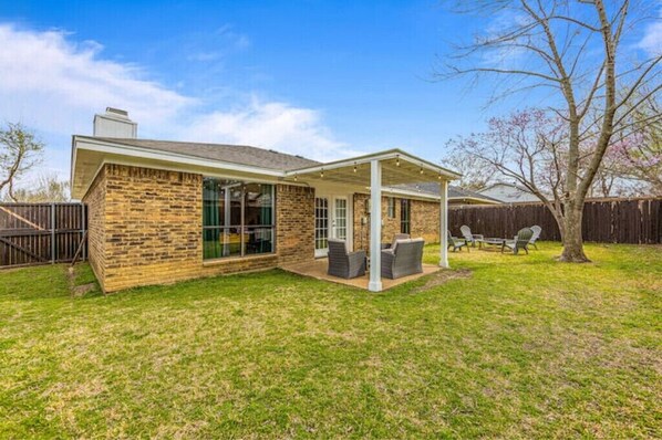 Une maison de plain-pied avec une façade en briques, un patio couvert aménagé avec du mobilier d’extérieur et une cour arrière clôturée.