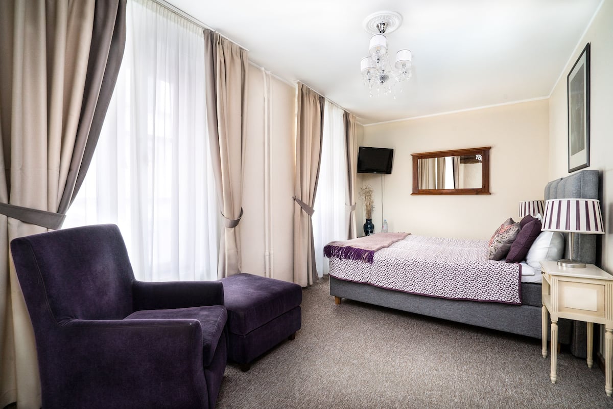 Quarto de hotel com cama, uma cadeira, uma mesinha, televisão e um lustre.