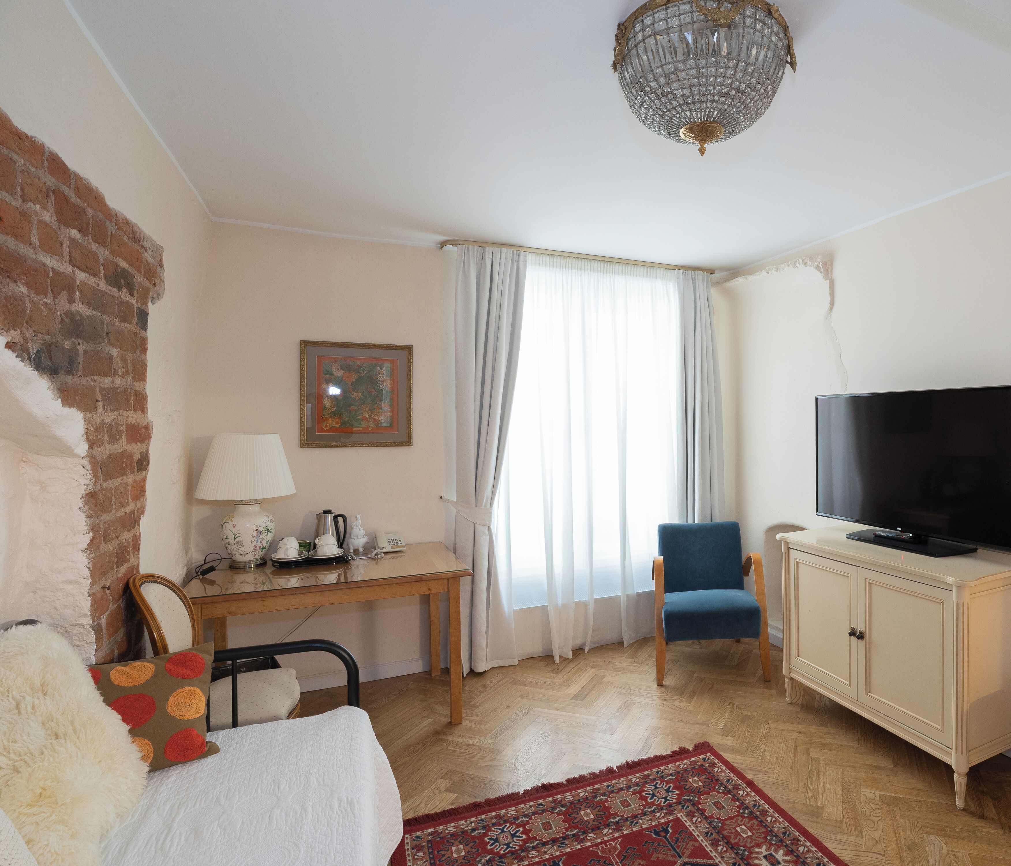 A room with a wooden desk, a blue chair, a television, a patterned rug, and a chandelier.
