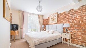 Une chambre avec un mur en briques, un lit avec une literie blanche, une table de chevet avec des lampes et un téléviseur posé sur un meuble en bois.