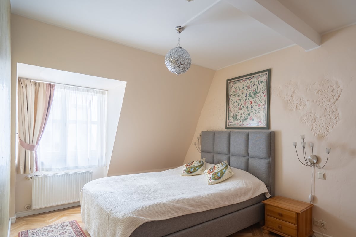 Um quarto com cama, mesa de cabeceira, radiador, janela com cortinas, lustre e luminária na parede.