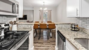 Une cuisine moderne avec des meubles blancs, un plan de travail en granit, une cuisinière noire et un coin repas agrémenté d’un lustre.