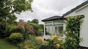 En trädgård med ett stort utbud av växter och blommor, en uteplats med utemöbler och ett hus med ett stort fönster.