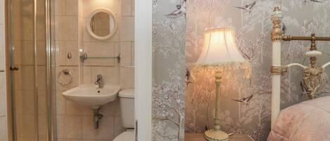 Un baño con cabina de ducha de vidrio, lavamanos e inodoro. Se observa una lámpara decorativa sobre una mesita de noche con un diseño de papel tapiz floral.