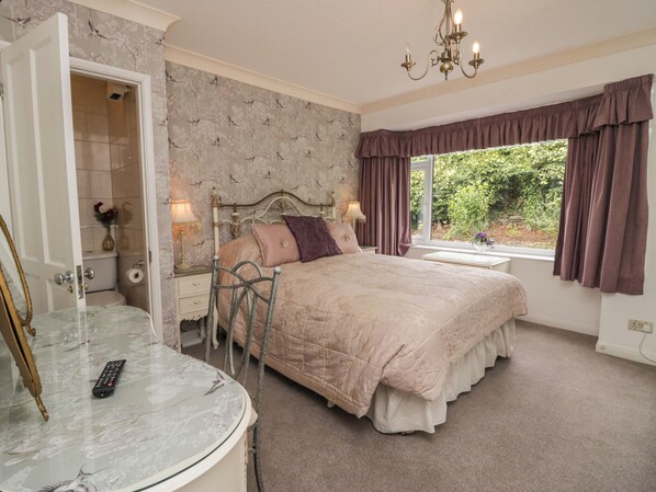 A bedroom with a large bed, a vanity with a mirror, a chandelier, and a view of greenery outside.
