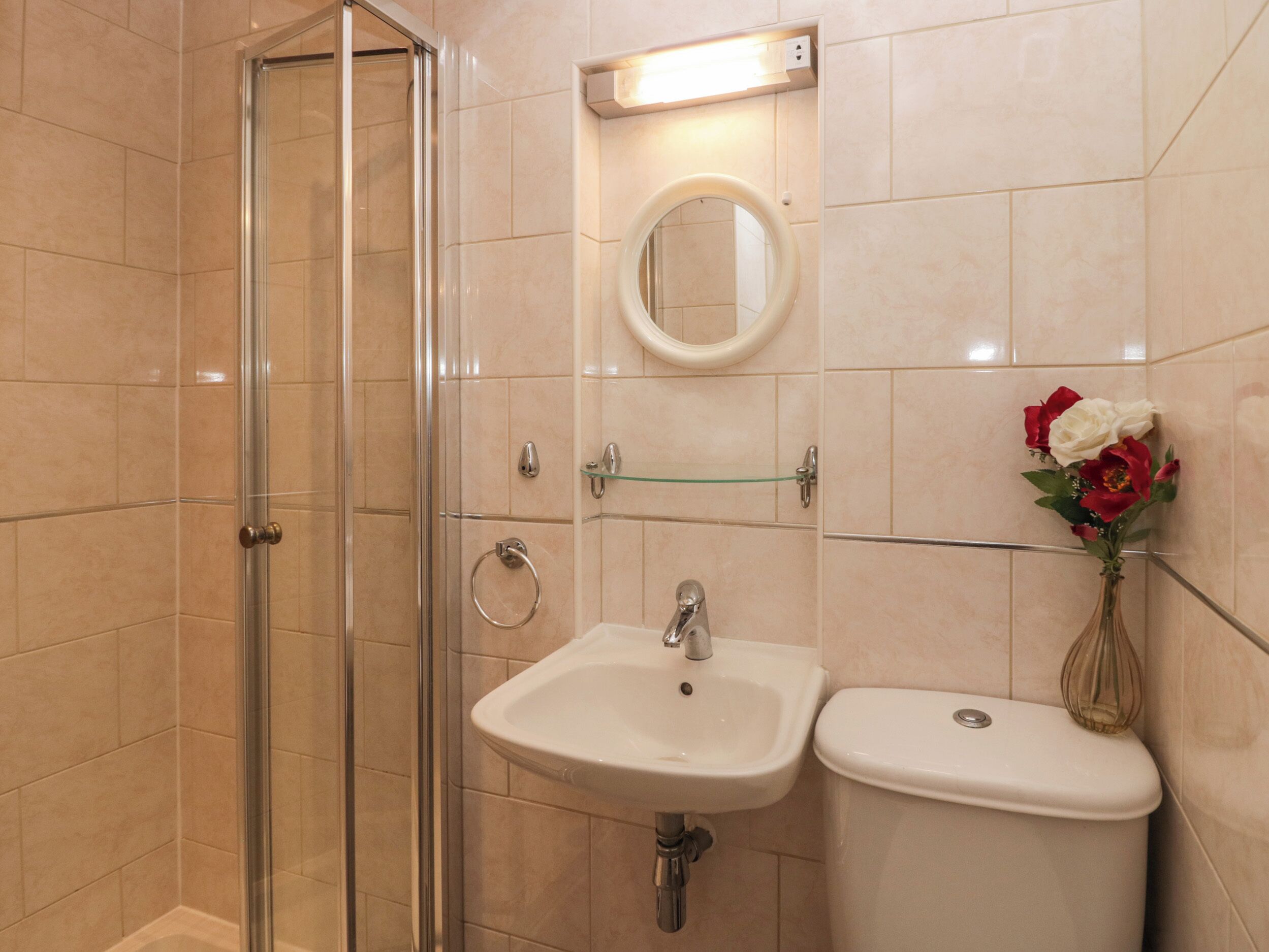 A bathroom with a glass shower enclosure, a sink, a toilet, a mirror, and a vase with flowers.