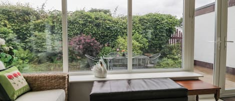 A room with a large window, a wicker sofa, a black ottoman, and a small wooden table.