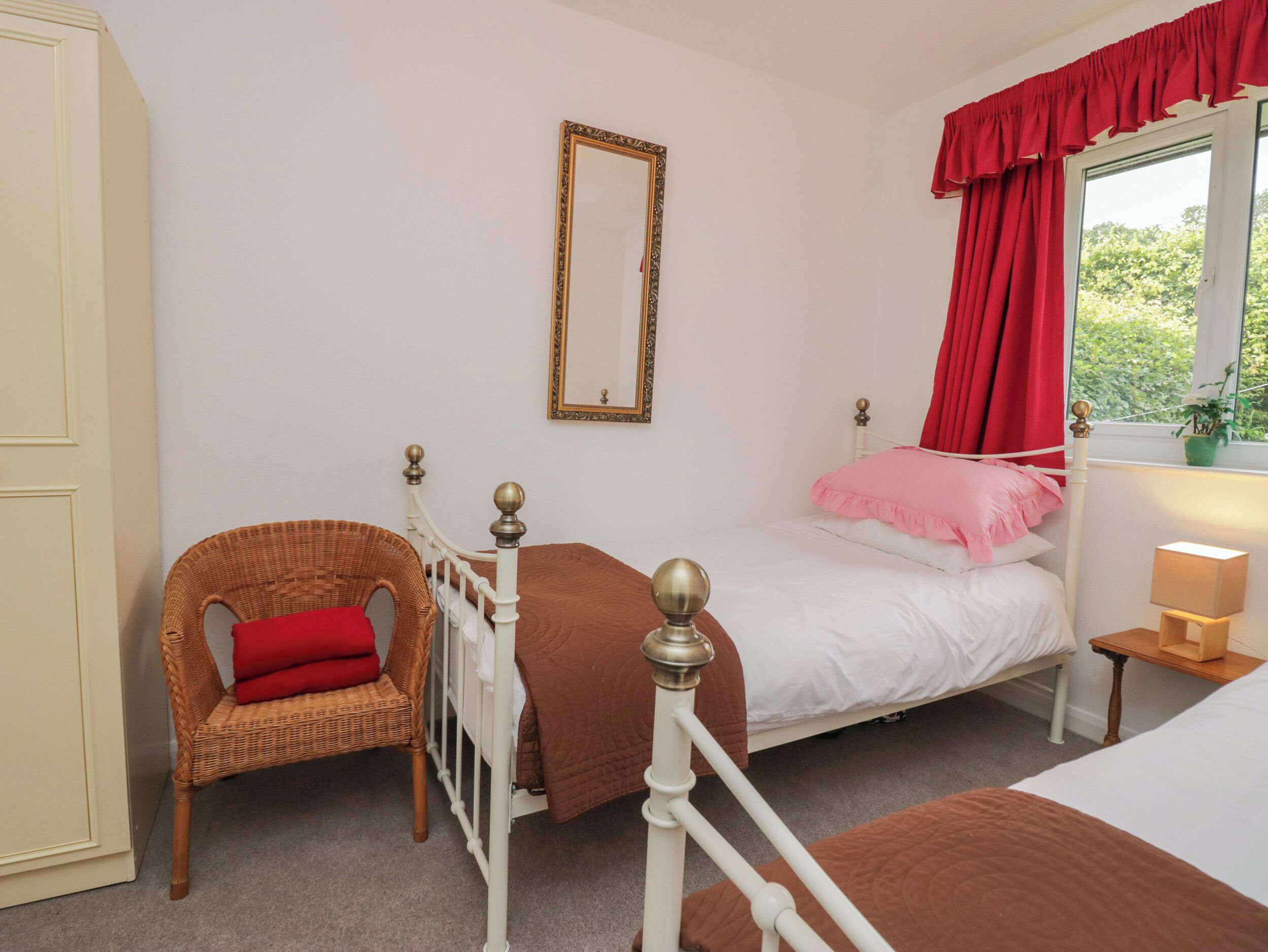 Un dormitorio con una cama individual, un sillón de mimbre con cojín rojo, una mesita de noche con lámpara y una ventana con cortinas rojas.