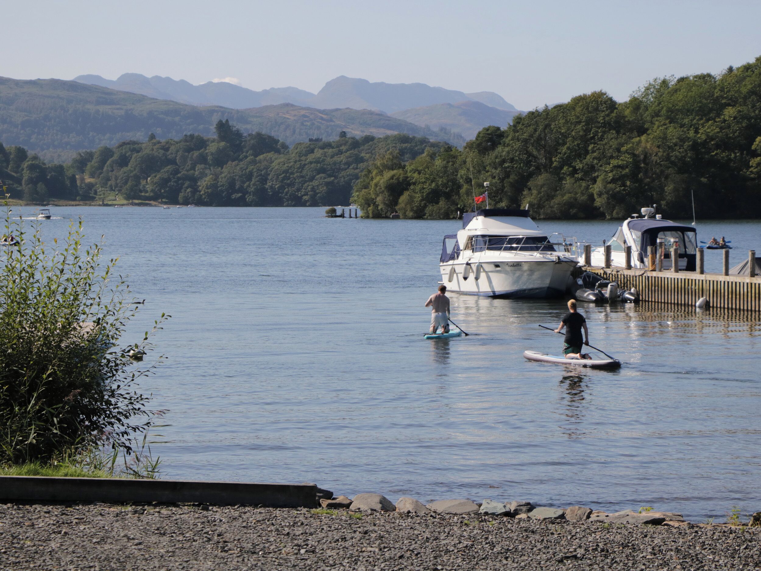 A calm lake with boats docked at a pier, surrounded by lush greenery and distant mountains.
