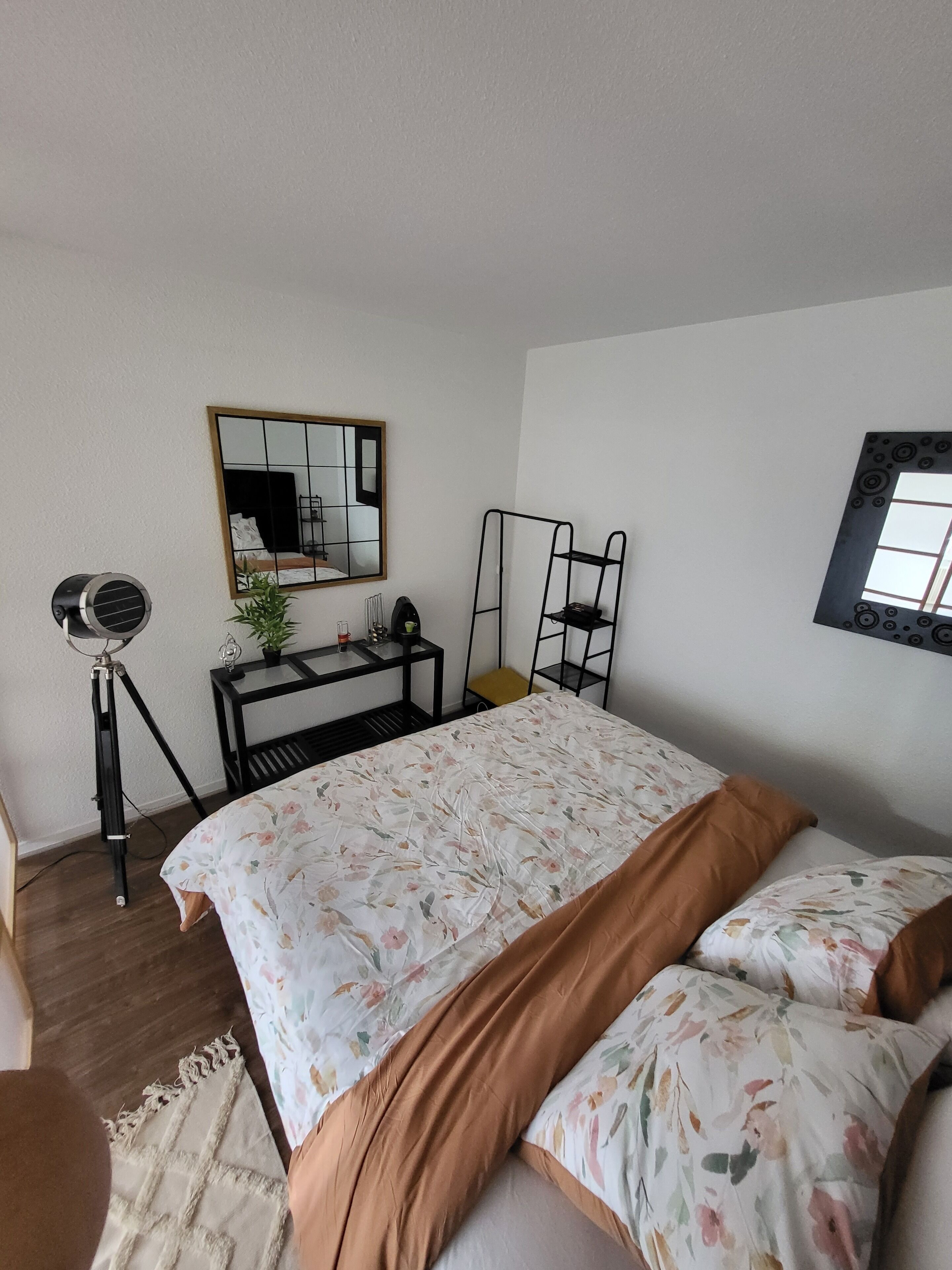 A bedroom with a bed, a desk, a chair, a tripod, and a mirror.