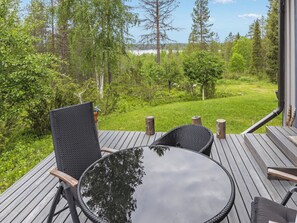 An outdoor deck with a round table and chairs, surrounded by a lush green landscape and trees. - Mustikka by Interhome (Kuusamo)