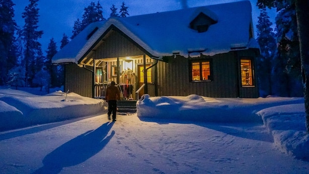 A wooden cabin with a snow-covered roof, a porch with steps leading to the entrance, and a person walking towards the door. - Mustikka by Interhome (Kuusamo)