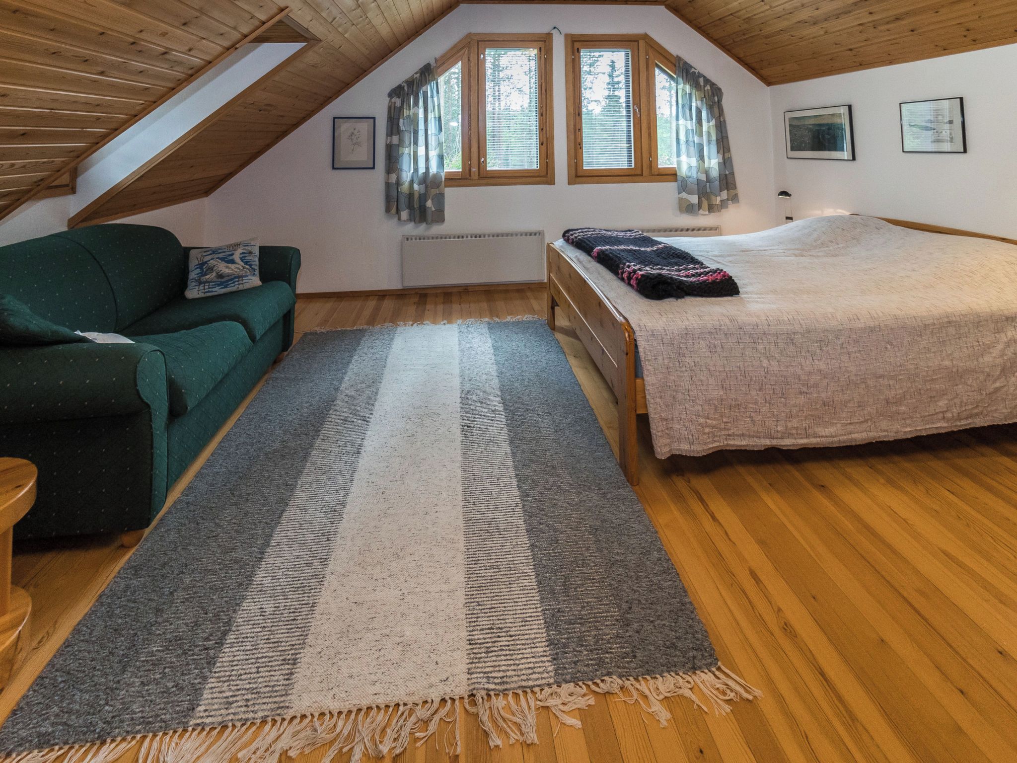 Una habitación con techo inclinado, una cama, un sofá, un ventanal con persianas y una alfombra.