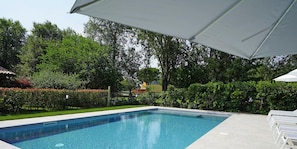 A rectangular swimming pool with clear blue water, surrounded by a well-maintained hedge and trees. There are white lounge chairs and a large umbrella providing shade. - Villa Caterina 3 - LOC (Desenzano del Garda)