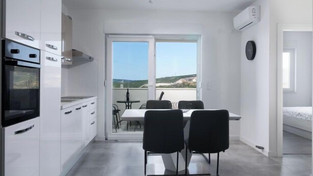 A modern kitchen with white cabinets and a built-in oven, a dining area with black chairs and a table, and a balcony with a view of the landscape. - Apartment Franka by Interhome (Trogir)