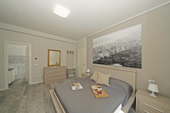 A bedroom with a bed, a nightstand, a dresser, a mirror, a door to a bathroom, and a cityscape painting on the wall.