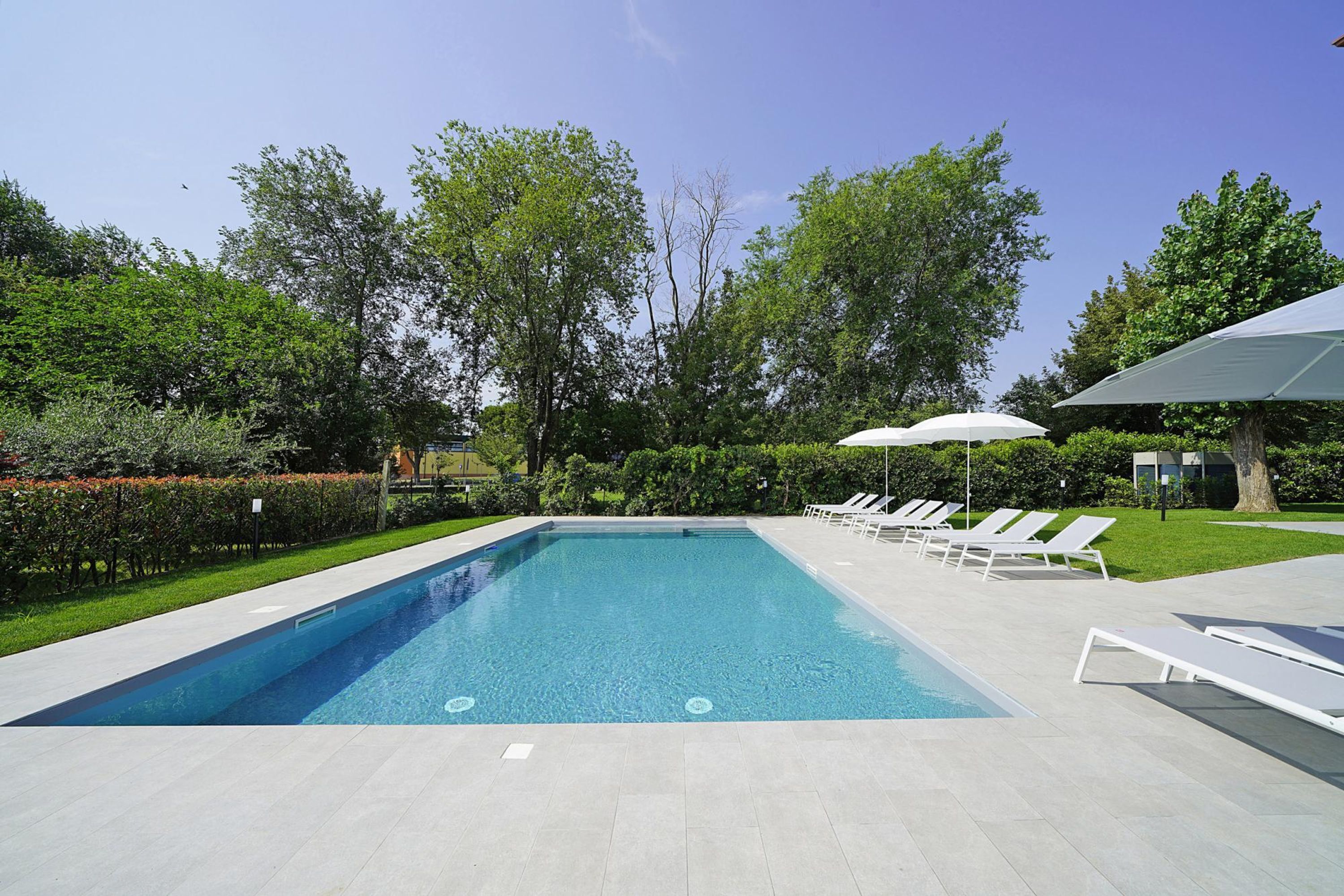 Une piscine rectangulaire, bordée d’une terrasse carrelée, entourée de chaises longues blanches et de parasols, et située dans un jardin aménagé avec des arbres et des haies.