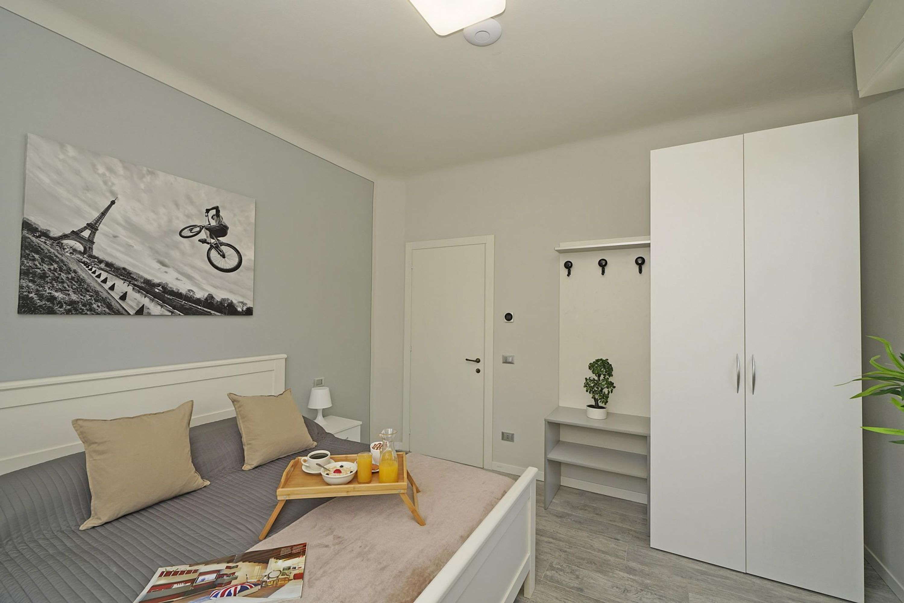 A bedroom with a bed, a bedside table with a lamp, a tray with breakfast items, a wardrobe, and a wall with a black and white photo.
