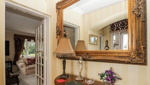 A room with a large ornate mirror, a wooden console table with a lamp, a statue, and a vase of flowers. - Storrs Lodge (Windermere)