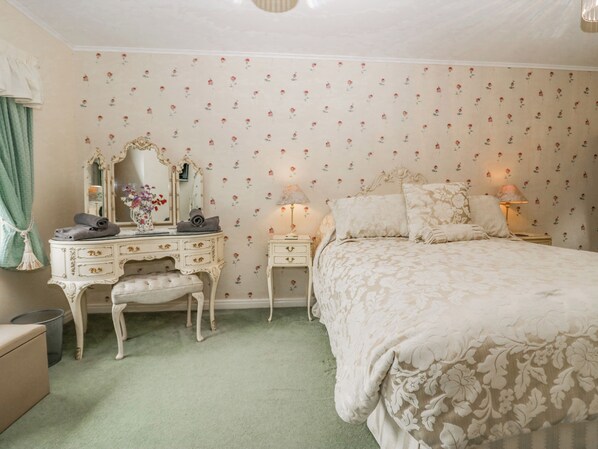 A bedroom with floral wallpaper, a vanity with a mirror, a bed with a floral bedspread, and two bedside lamps. - Storrs Lodge (Windermere)