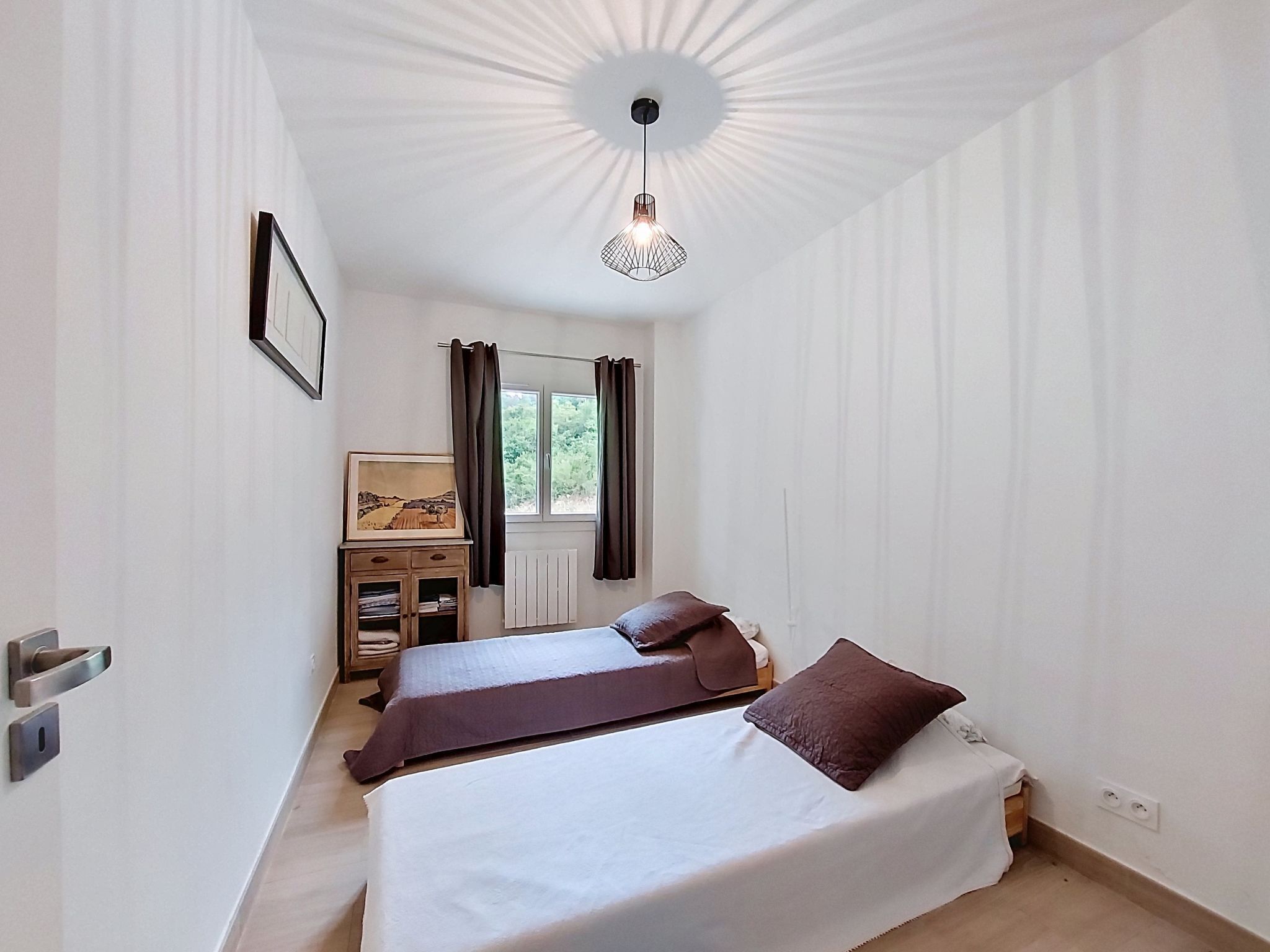 A bedroom with two beds, a window with curtains, a radiator, a framed picture on a cabinet, and a pendant light.