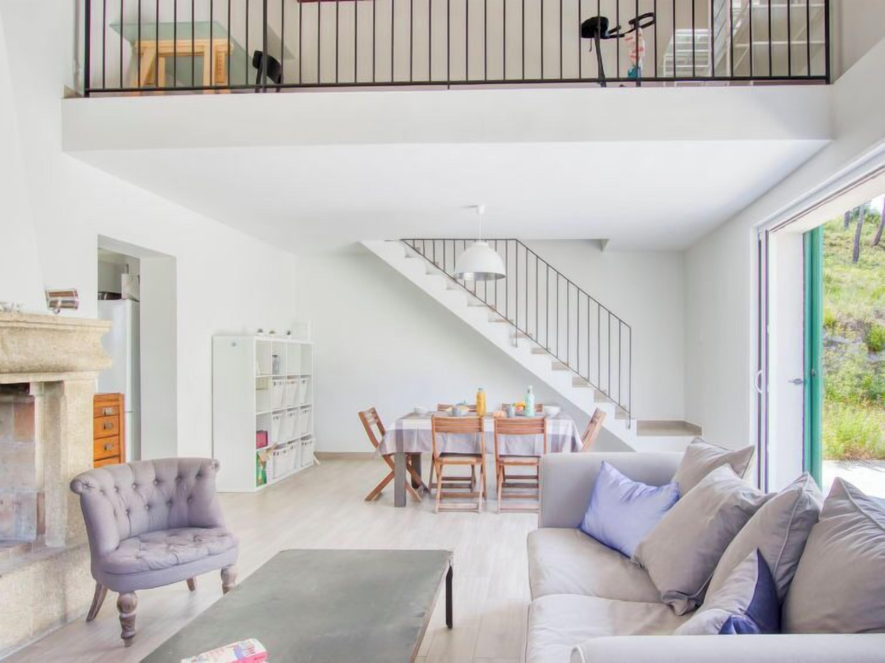 Un vaste espace de vie doté d’un haut plafond, une table à manger pour quatre personnes, un escalier et une grande fenêtre offrant une vue sur la verdure.