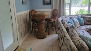 Ein Zimmer mit Esstisch und Stühlen, einer Couch mit gemusterten Kissen sowie Blick durch ein Fenster.