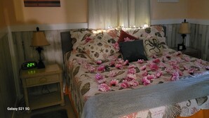 Ein ordentlich bezogenes Bett mit floraler Bettwäsche und rosa Blumen, eine Nachttischlampe, eine Uhr und ein Nachttisch.