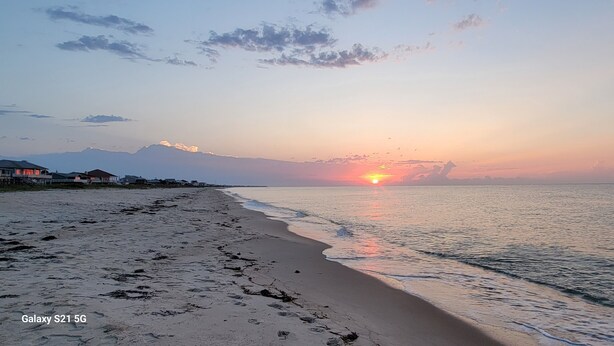 Ein Strand mit Fußspuren, die zum Wasser führen, ein Sonnenuntergang im Hintergrund und Häuser in der Ferne.